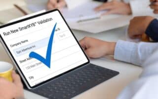 Person using a tablet to run a SmartKYB™ business validation for "Epic Adventure Inc" with a large blue check mark confirming successful verification.