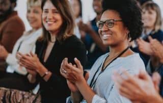 A diverse group of professionals smiling and applauding during a workplace event or presentation, conveying appreciation, recognition, and positive company culture.