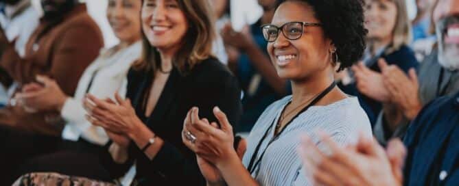 A diverse group of professionals smiling and applauding during a workplace event or presentation, conveying appreciation, recognition, and positive company culture.