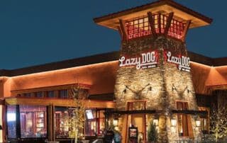 Exterior view of a Lazy Dog Restaurant at night, featuring warm lighting, rustic stone architecture, and the Lazy Dog logo prominently displayed on the tower.